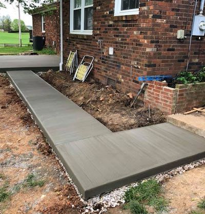 Newly poured concrete sidewalk beside a brick house, partially framed by soil and gravel.