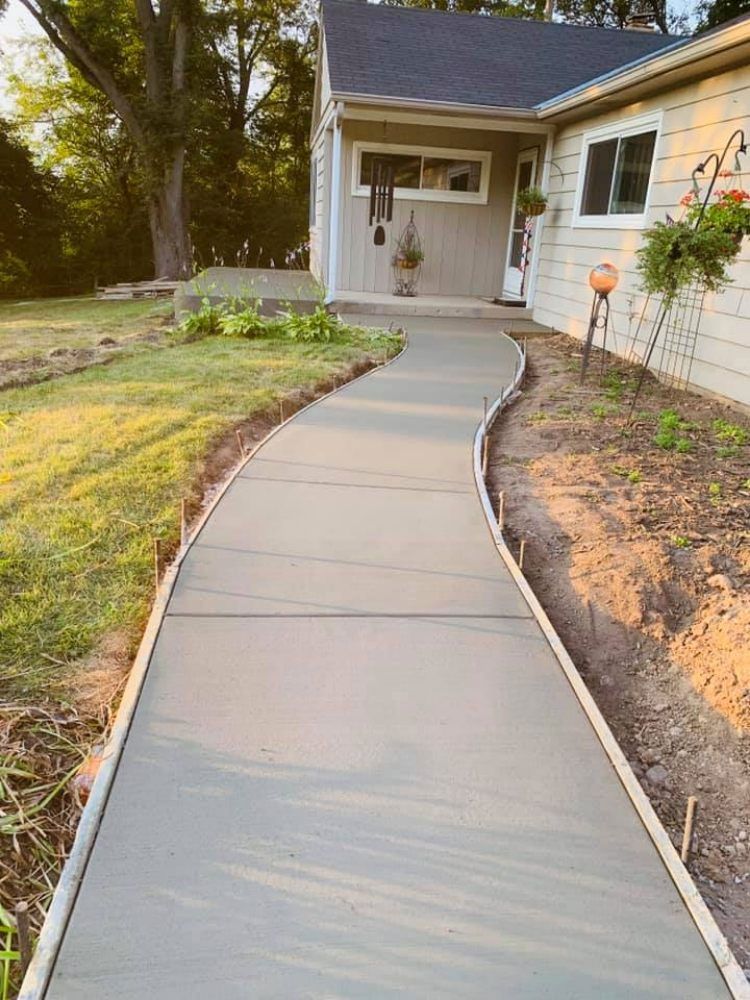 A newly poured concrete walkway curves toward a house, bordered by wooden forms and a grassy yard.