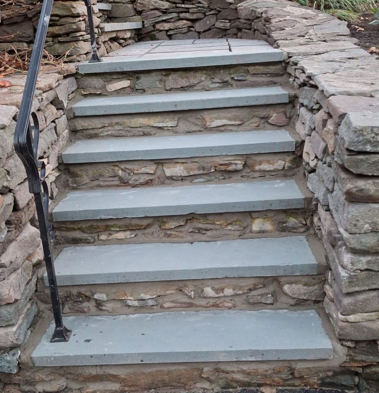 Stone steps with a black railing, built into a stone wall.