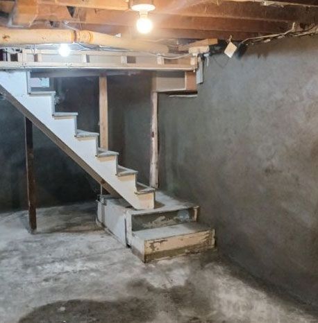 Basement with white stairs, gray walls, and concrete floor. Two lights hang from wood ceiling.