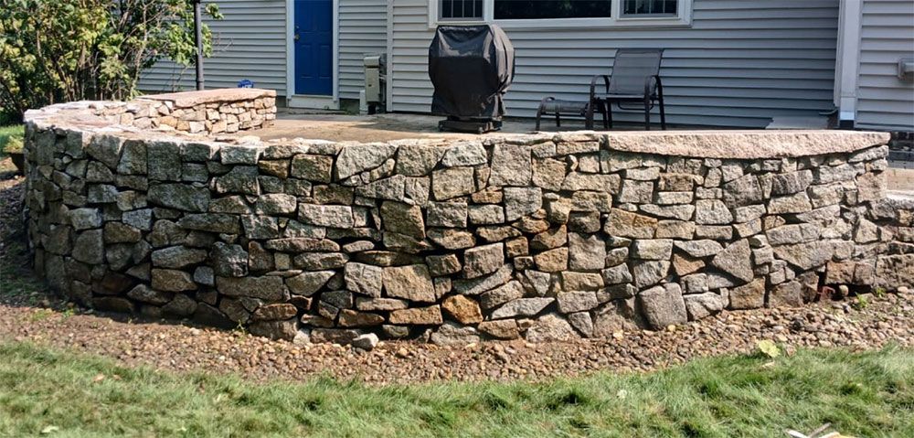 Stone retaining wall encloses a patio area with a grill and chair. Green grass in the foreground, house in the background.