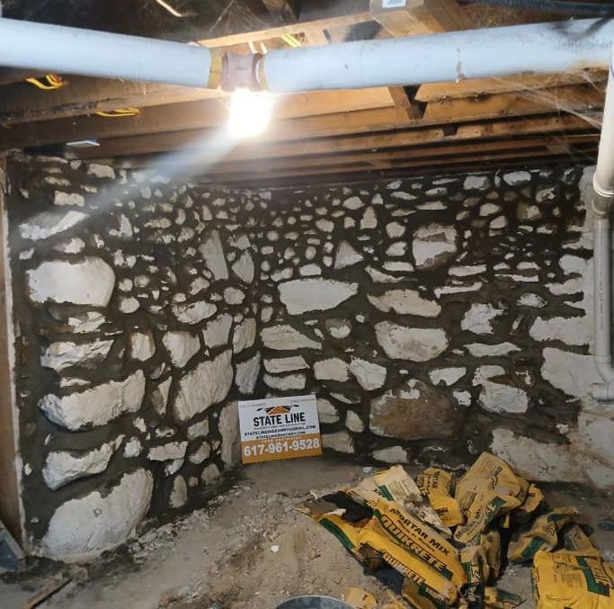 Stone basement wall under construction with mortar. Construction materials and pipes overhead.