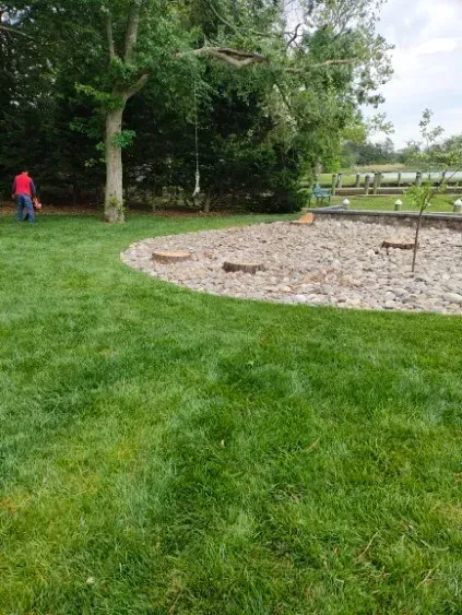 Lawn with a person mowing near a stone garden bed and a tree. Lush green grass and a bright day.