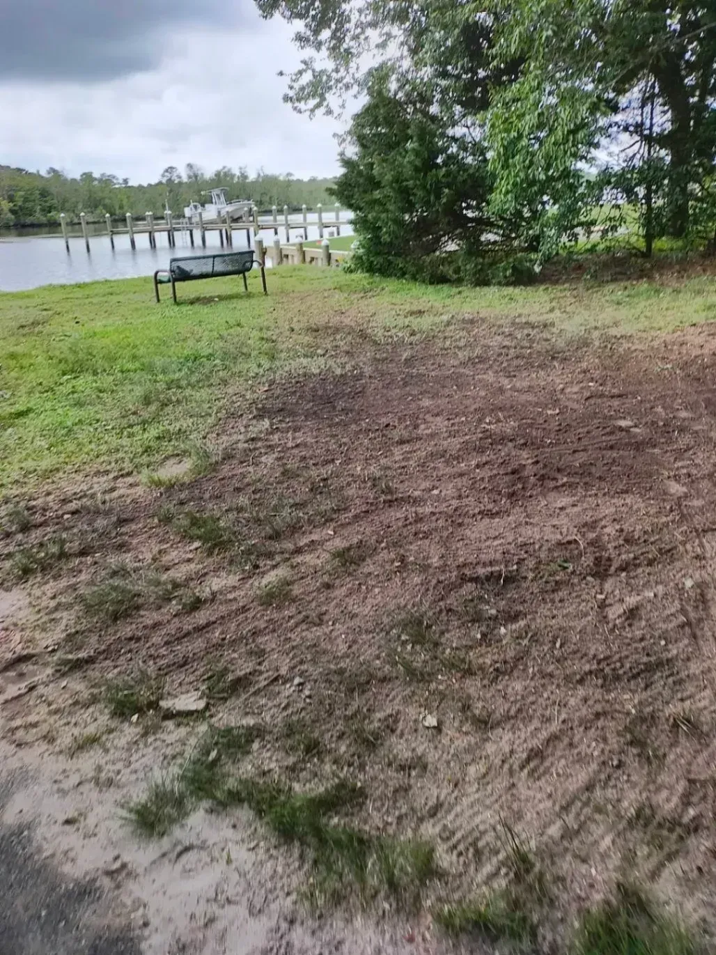 Grassy area with a dark patch of dirt near a waterfront dock and trees under an overcast sky.