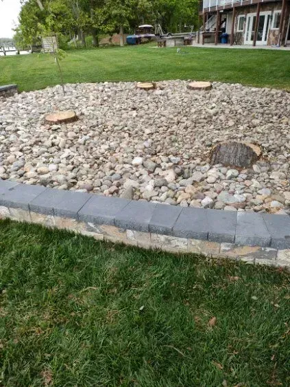 Stone bed with tree stumps, edged by a brick wall and grass, near a building and greenery.