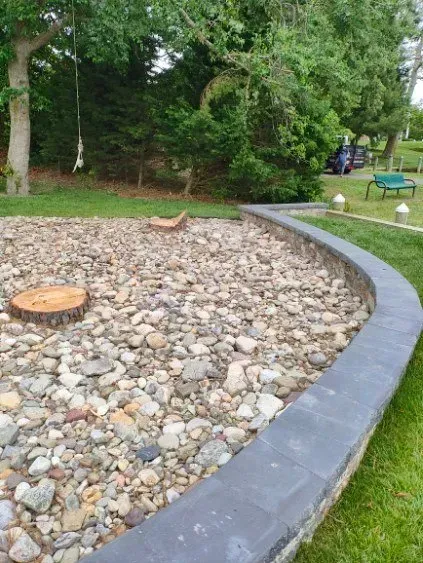 Gravel-filled garden bed with stone edging, tree stump, and surrounding grass.