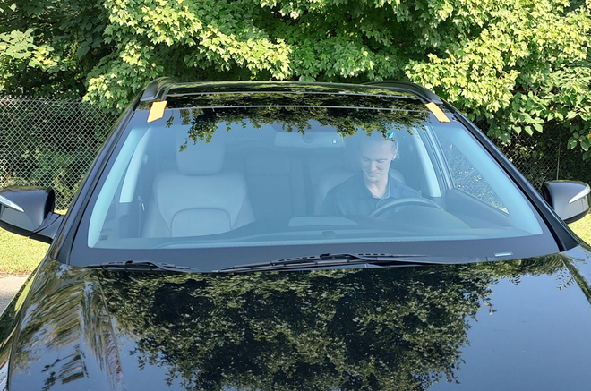 Person sitting in a black car, seen through the windshield, parked among green trees.
