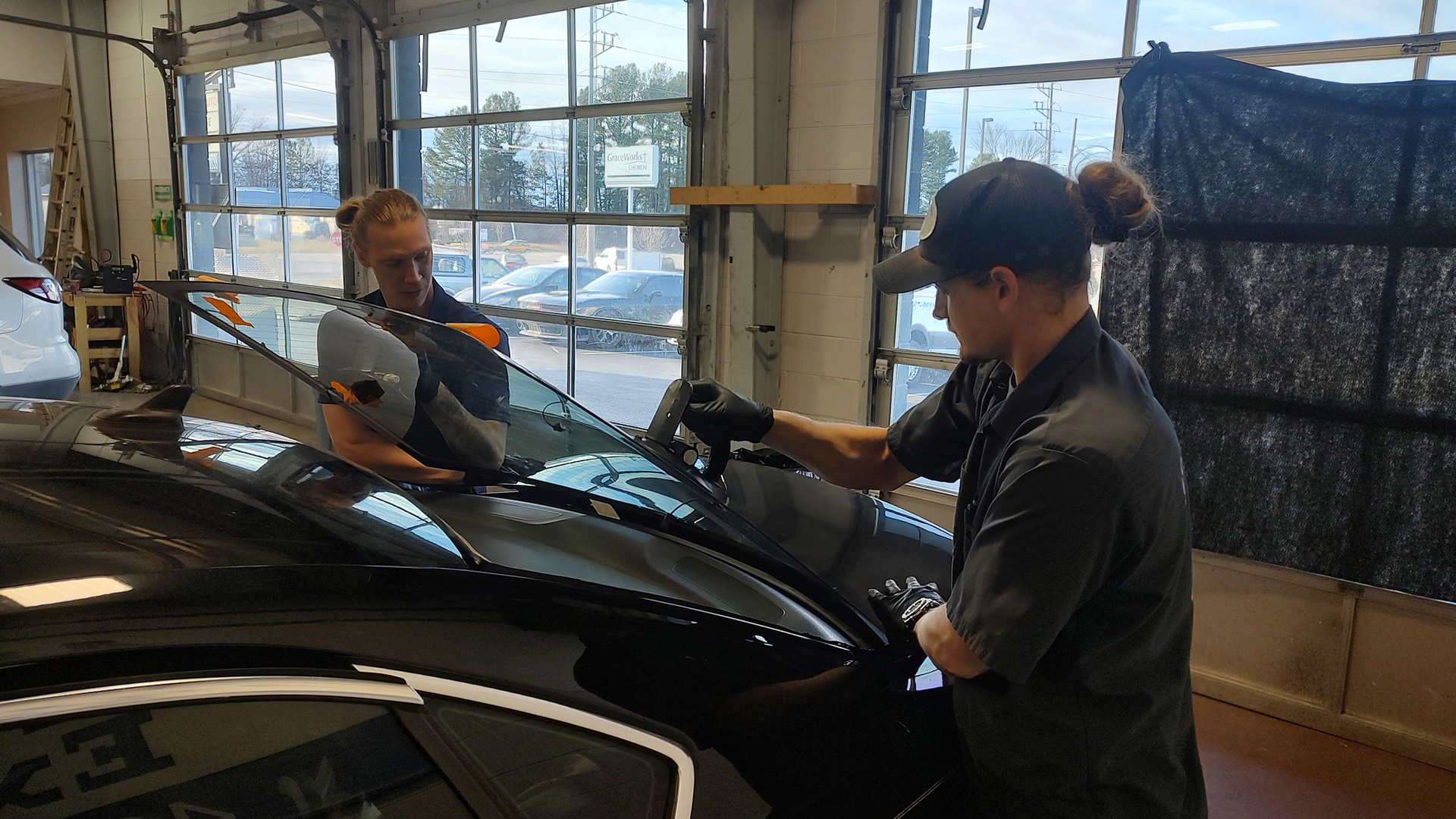 Two mechanics inspect a black car inside a garage, with large windows and equipment in the background.