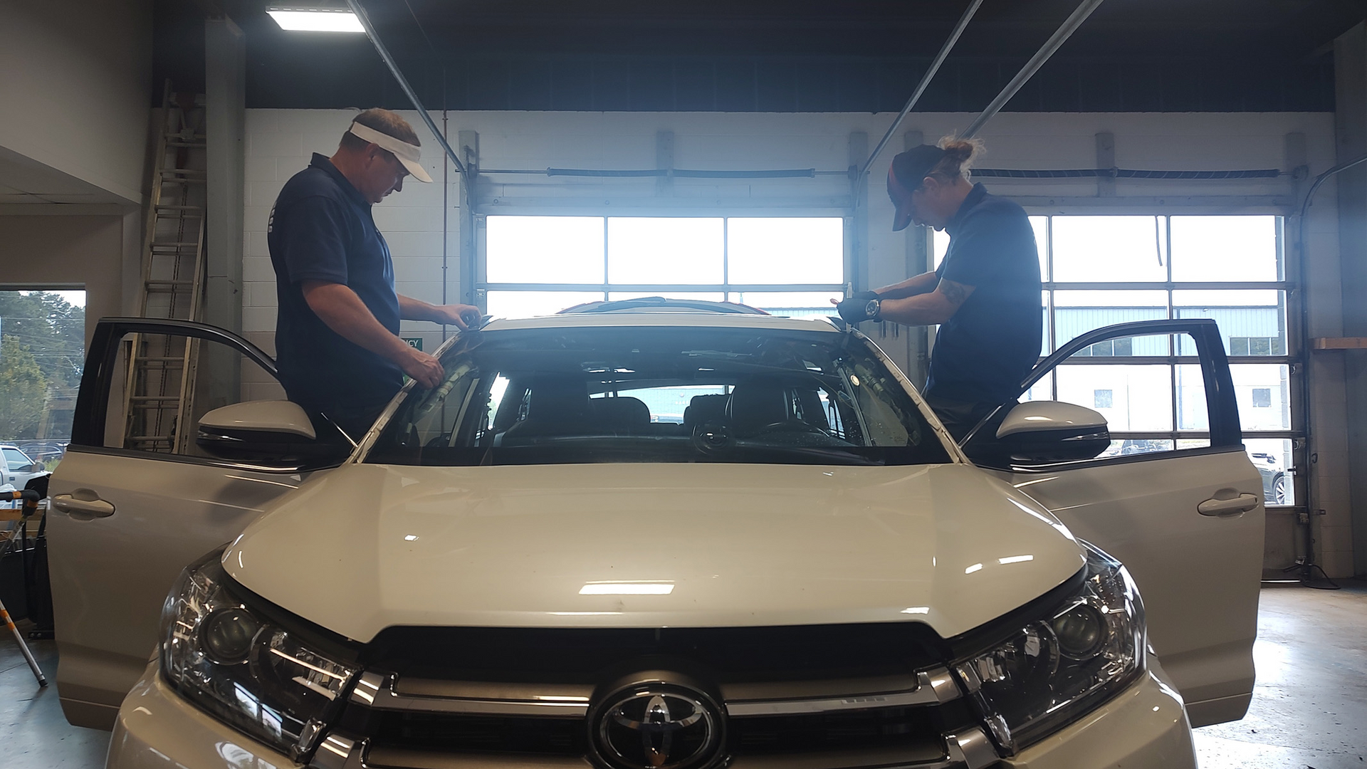 Two mechanics work on a white Toyota inside an auto garage with both front doors open.