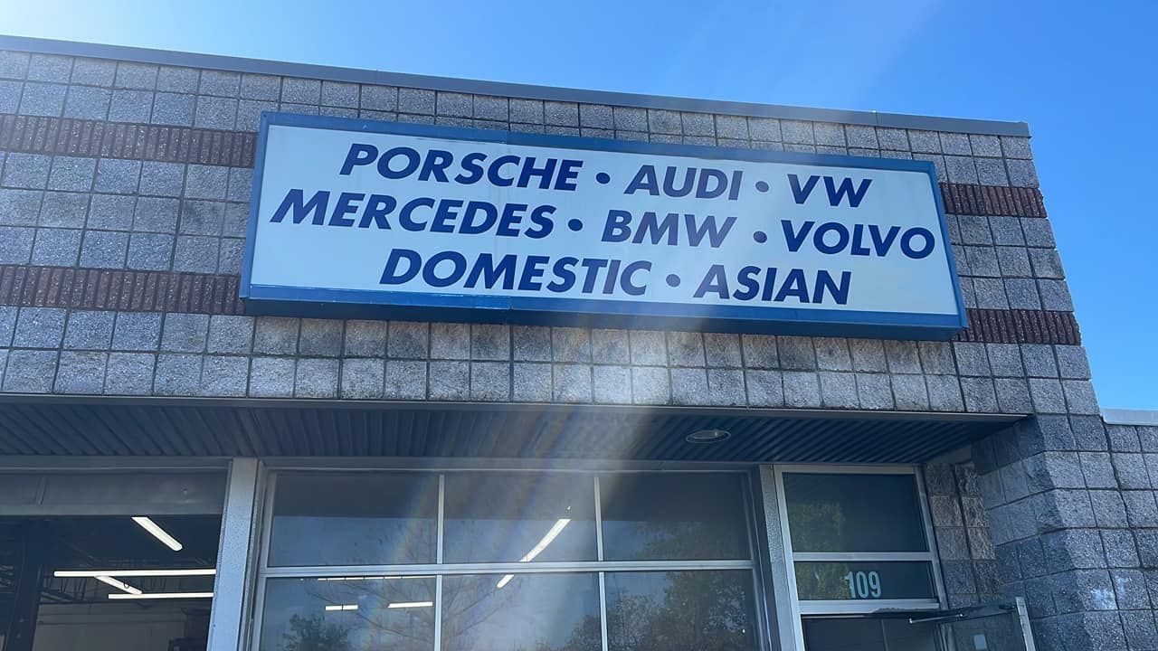 Sign on a brick building lists car brands: Porsche, Audi, VW, Mercedes, BMW, Volvo, Domestic, Asian.
