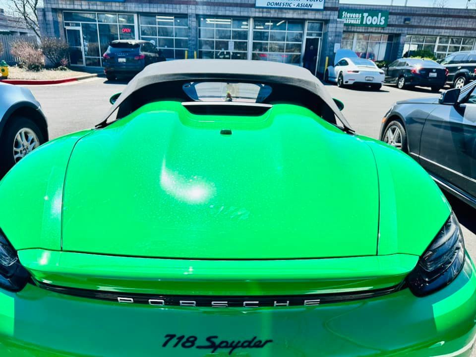 Bright green Porsche 718 Spyder convertible, parked in a lot, with shop in background.