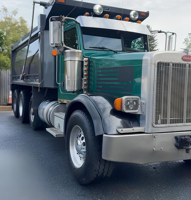 A dark green dump truck with a metallic grille and bumper parked outdoors on a paved lot.