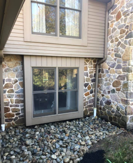 A ground-level view of a house exterior featuring tan siding, a window, and a foundation made of multi-colored fieldstone.