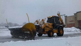 Yellow backhoe loader with a large snowplow blade clearing a snowy parking lot next to a building.