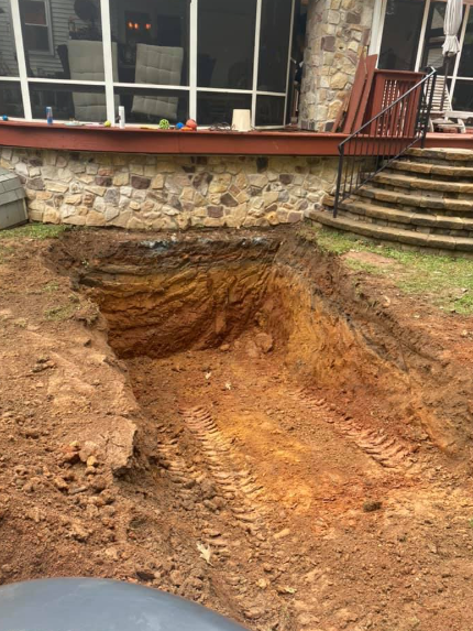 A deep, rectangular excavation in a grassy backyard, located in front of a stone-foundation house with a deck and stairs.