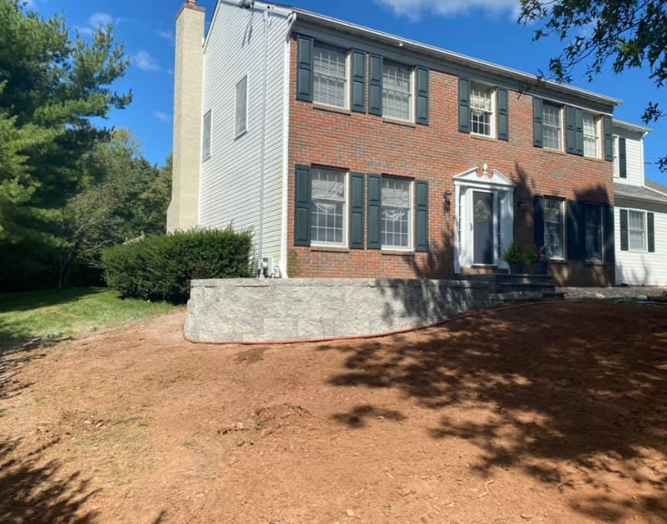 A two-story brick colonial home with a light-colored stone retaining wall in the front yard and a bare, graded lawn.