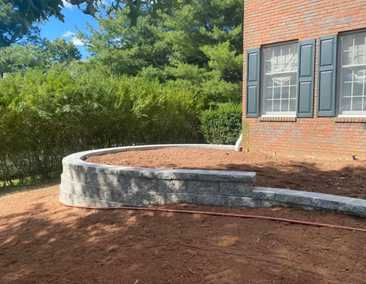 A curved stone retaining wall newly installed in front of a brick house with dark shutters and surrounding greenery.