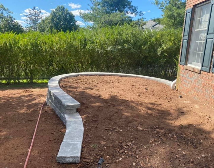 A curved stone retaining wall under construction in a dirt backyard next to a brick house with green shutters.