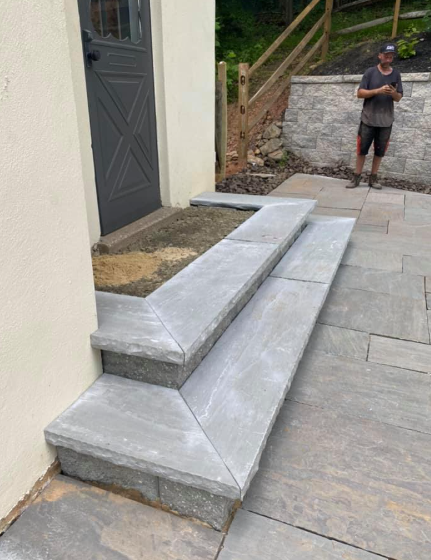 Two grey stone steps leading to a dark door, set beside a patio with a person standing in the background.