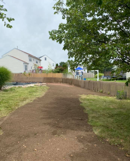A patch of freshly tilled, bare soil curving through a backyard lawn with a wooden fence and houses in the background.