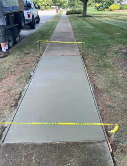 A freshly poured, smooth concrete sidewalk segment sectioned off with yellow caution tape in a suburban neighborhood.