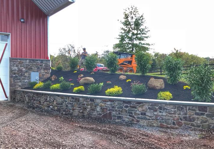 A stone retaining wall sits in front of a sloped landscape with mulch, decorative rocks, and shrubs beside a red building.