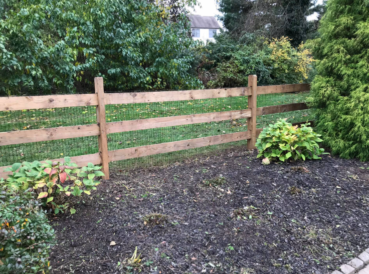 A wooden three-rail fence borders a garden bed with mulch and green shrubs in a grassy backyard.