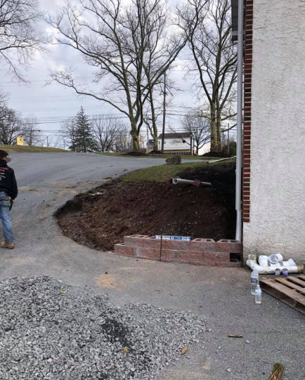 A worker stands near a low brick retaining wall being built against the side of a building near an asphalt driveway.
