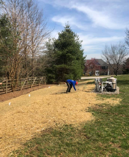 A person in a blue jacket works in a yard covered with straw, near pool equipment and a wooden fence.