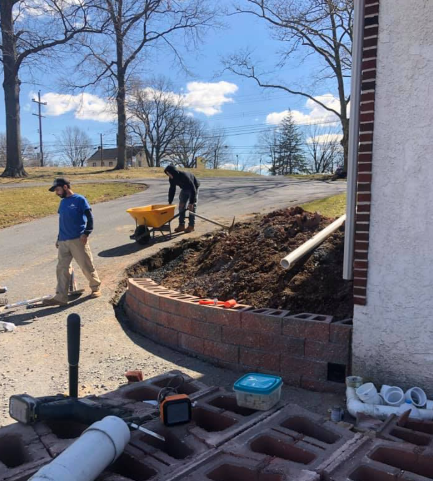 Two workers build a small, red stone retaining wall in a yard, with soil, construction tools, and pipes nearby.