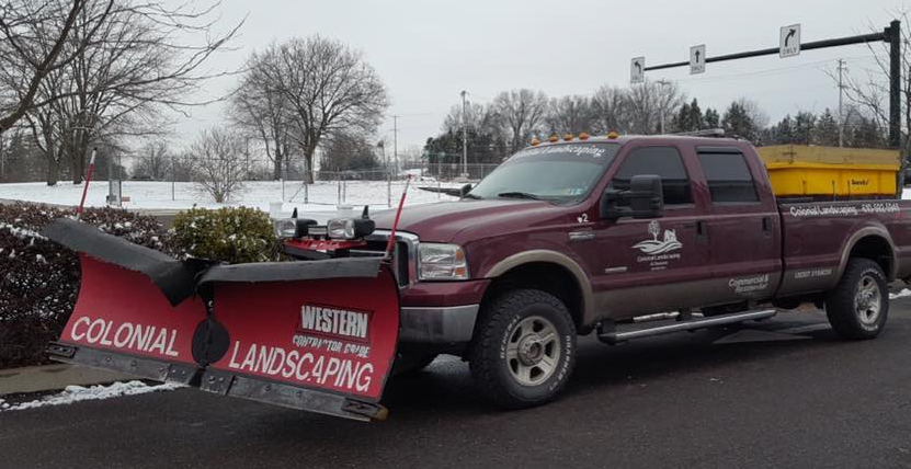 A maroon pickup truck with a large red Western brand snowplow attached to the front, parked in a snow-dusted lot.