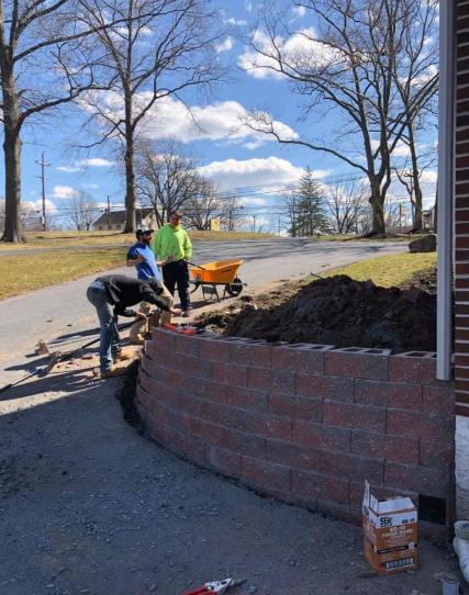 Three construction workers build a curved retaining wall with stacked blocks near a brick building.
