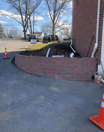 A new red brick retaining wall curves along a paved driveway next to a brick building with several drainage pipes.