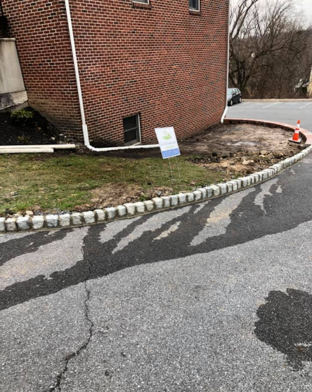 A brick building corner with a downspout, a basement window, a lawn, stone edging, and a sign beside a paved driveway.