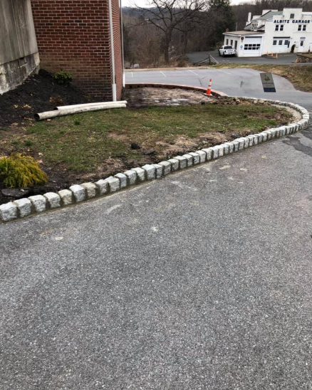A curved cobblestone border edges a grassy patch next to a brick building and a paved driveway.