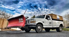 A white pickup truck with a large red snow plow attached to the front, parked on asphalt under a cloudy sky.