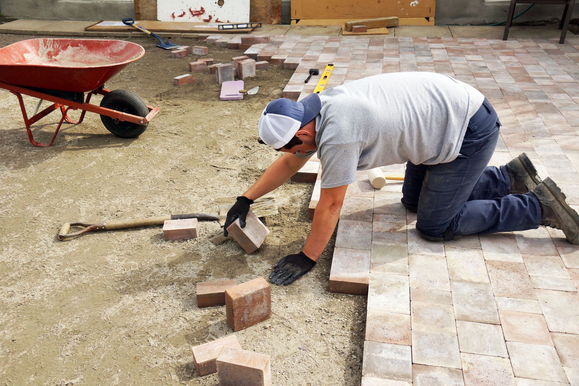 Man kneeling, laying brick pavers on a patio; wheelbarrow and tools visible.