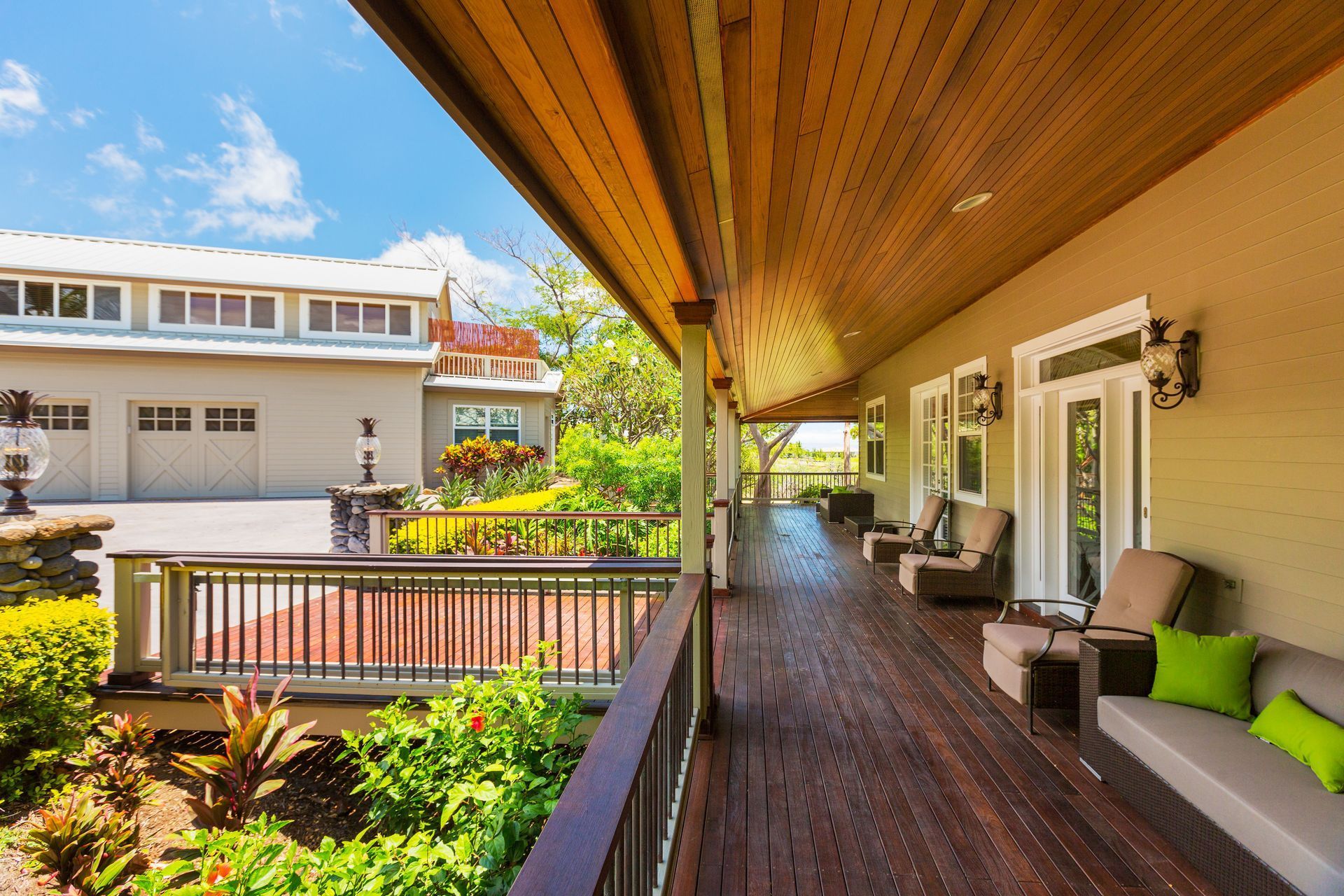 Long wooden porch of a house with seating, overlooking lush greenery, and a building on a bright day.