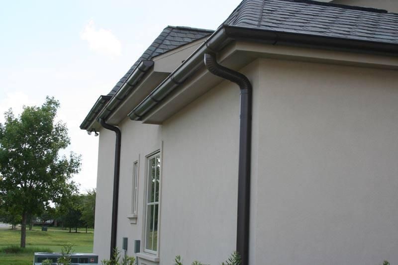 Brown gutters on a light-colored stucco house. Dark roof and window visible; green lawn background.