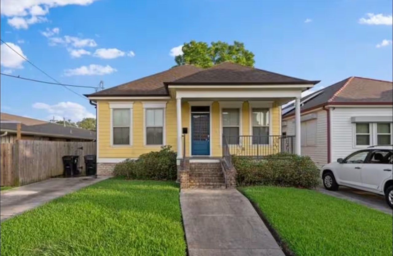 Yellow house with blue door, porch, and a sidewalk leading to it. Green grass and blue sky.