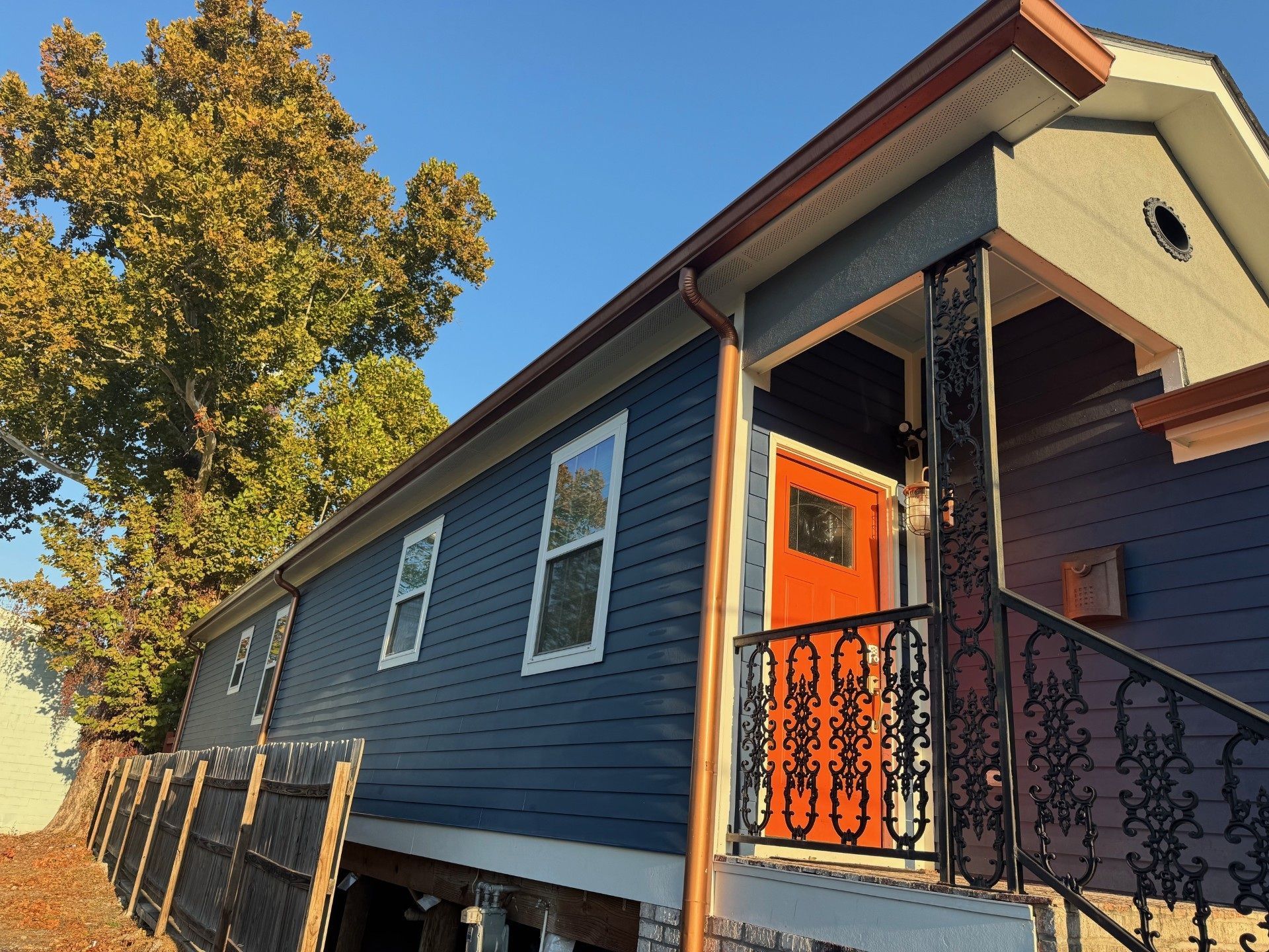 Blue house with orange door, black railing, and copper gutters under a clear sky.