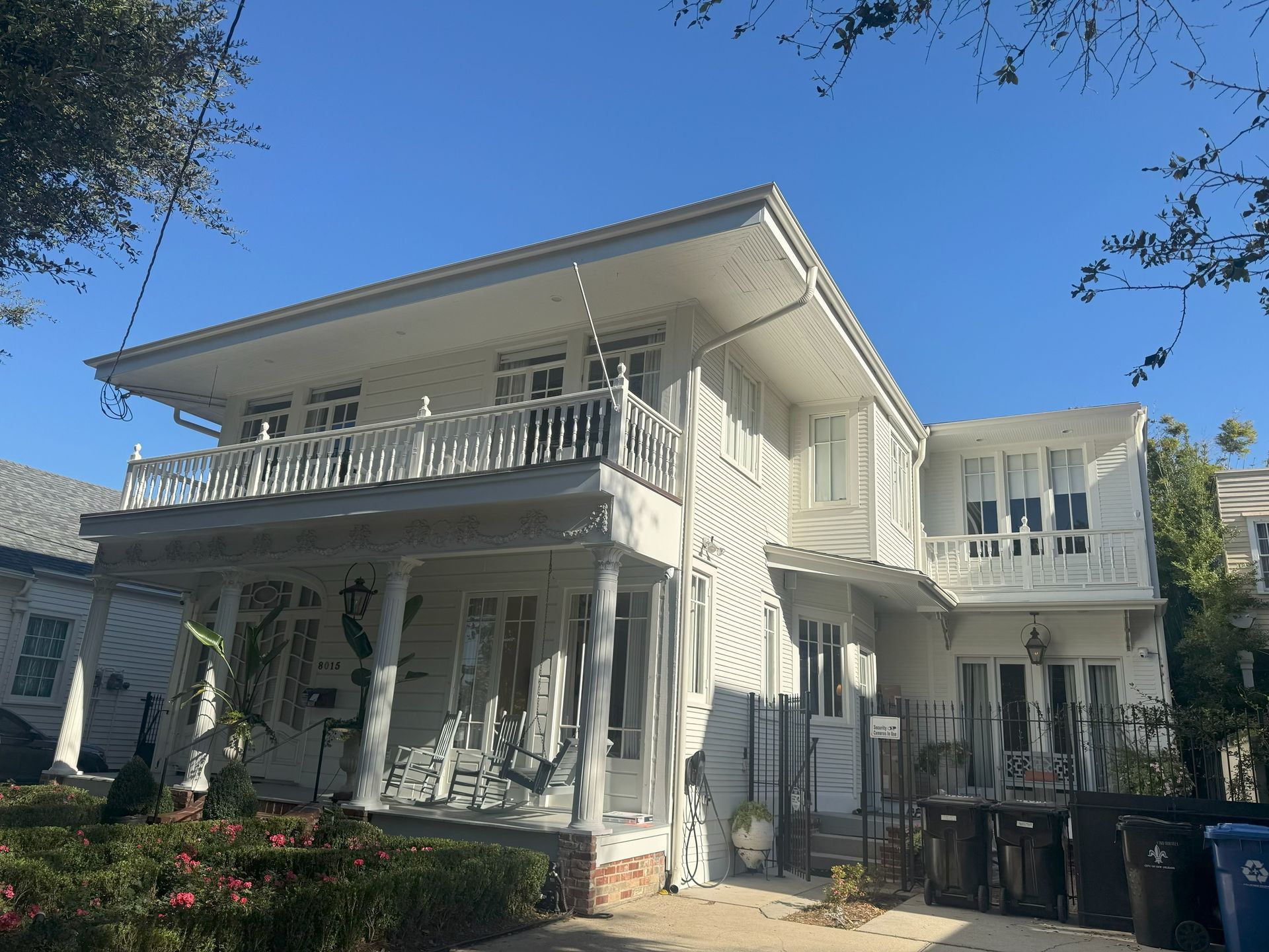 Two-story white house with balcony, railings, and weathered exterior; sunny day.