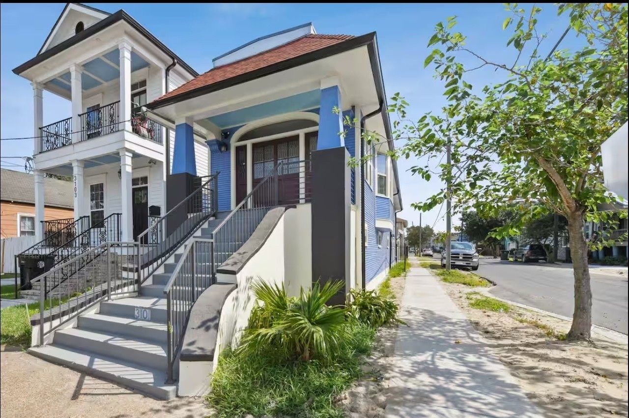 A blue and white two-story house with a front porch and steps, on a sidewalk, next to a street with a tree.