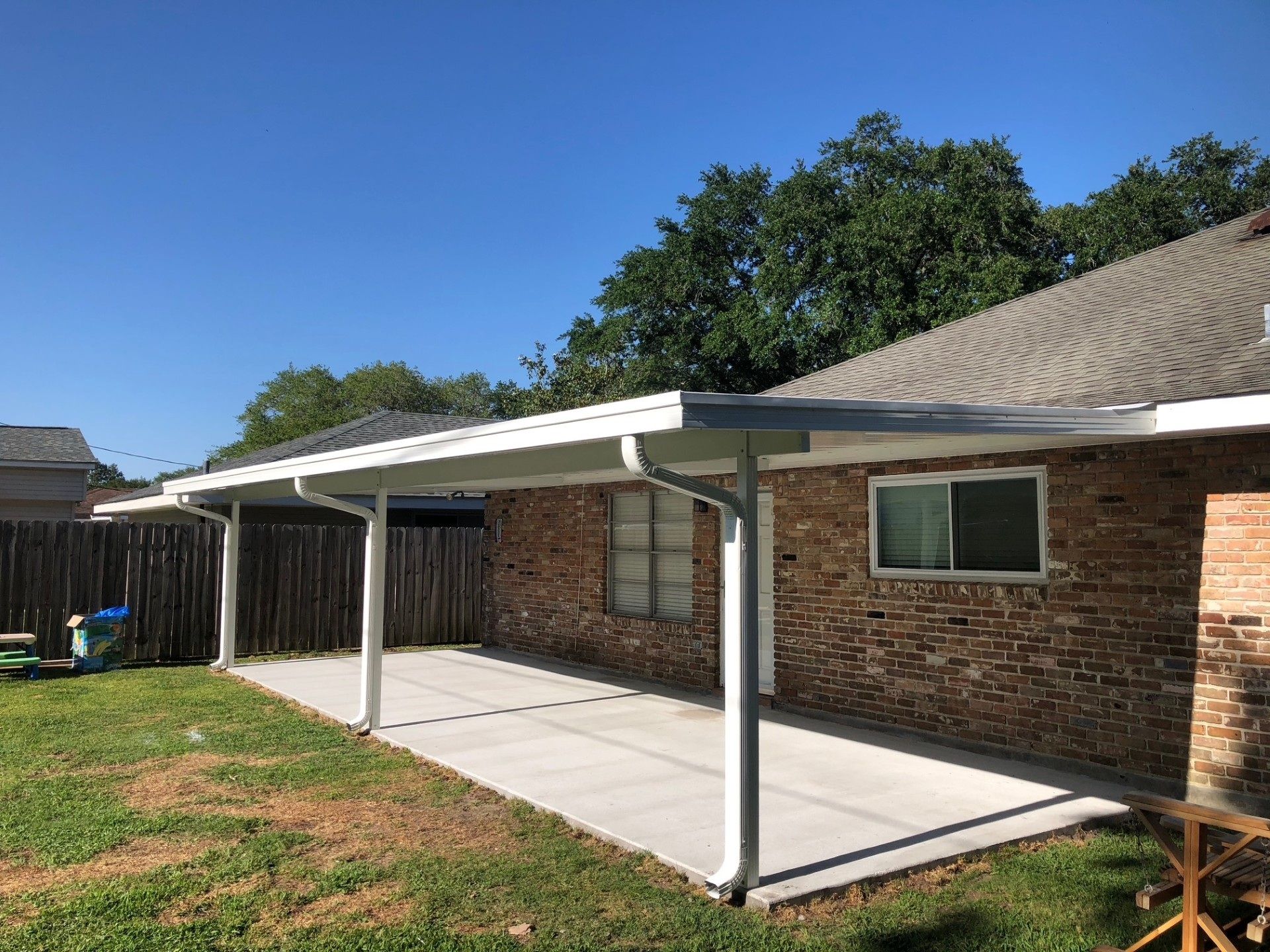 Patio with concrete floor and white supports, attached to brick house with a blue sky.