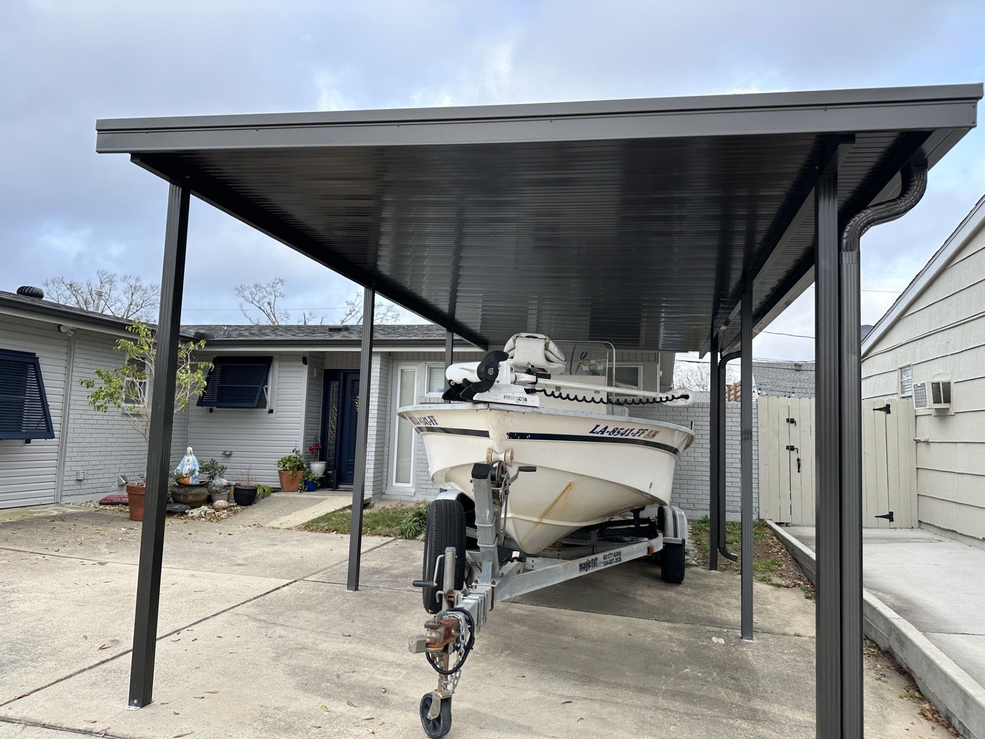 Boat on a trailer under a dark gray carport, in front of a house.