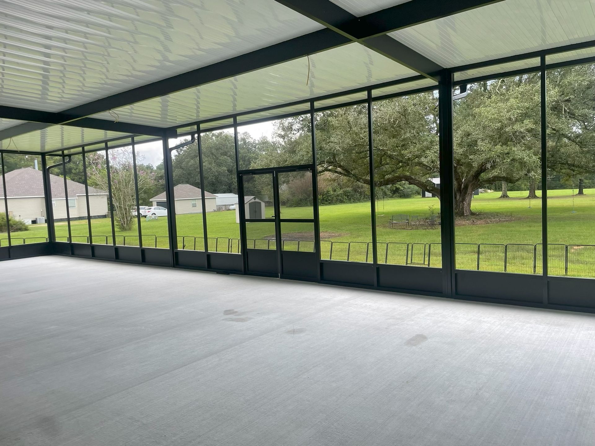 Covered outdoor patio with a concrete floor and glass walls, overlooking a grassy yard with trees.