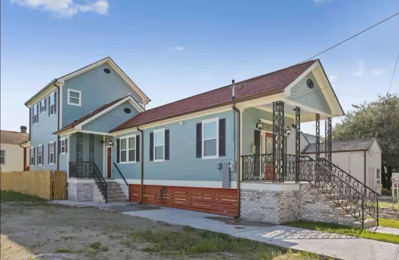 Two-story light blue house with a covered porch and red roof. Brick and stone foundation. Concrete walkway in front.