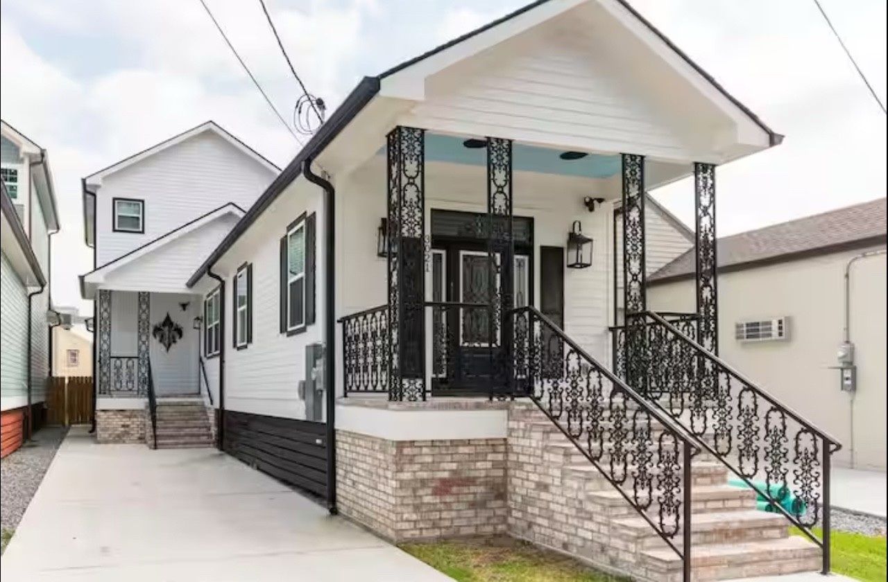 White house with black accents, brick base, and decorative ironwork on porch and stairs.