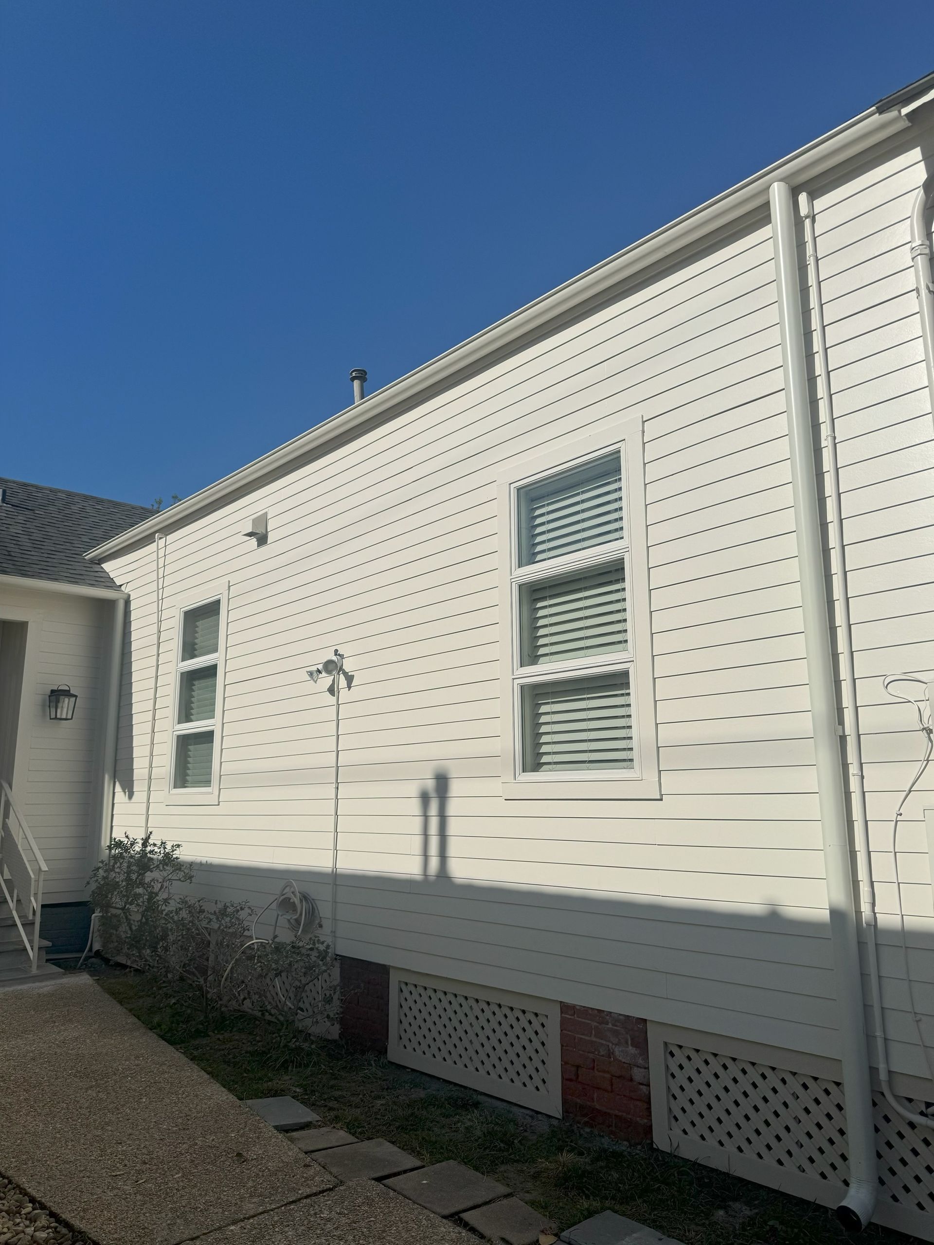 White siding of a house with two windows and a gutter, against a blue sky.