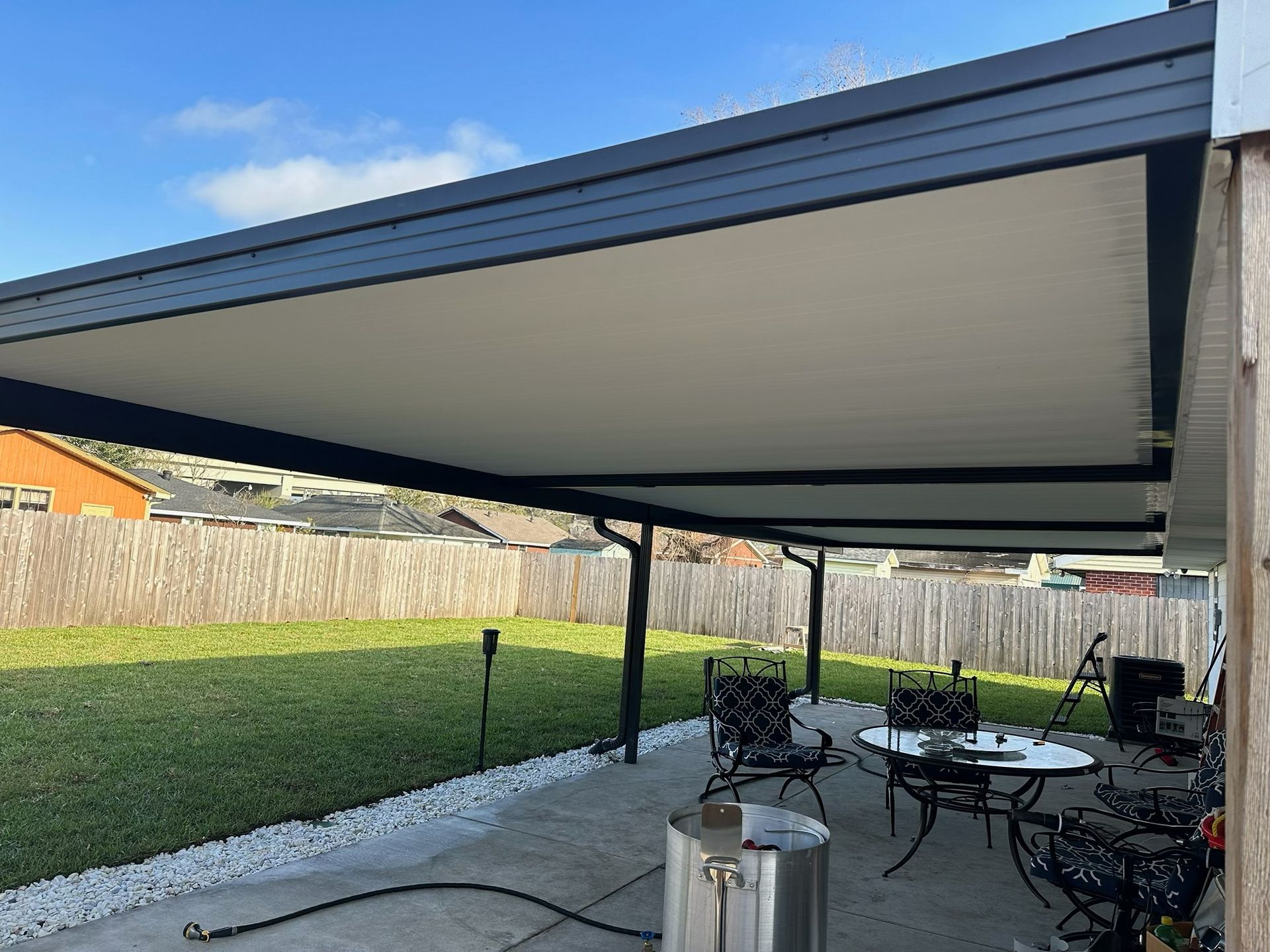 Patio with black awning, table and chairs on concrete, and lawn with fence in backyard.
